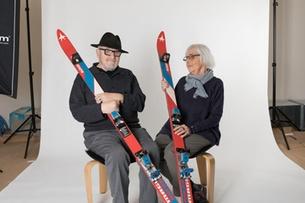 An elderly couple hold a pair of skis while sitting.